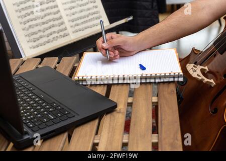 Detail eines jungen Mannes, der Notizen in einem Notizbuch gemacht hat Mit einem Computer auf einem tragbaren Holztisch mit Blech Musik im Hintergrund Konzept online Musik st Stockfoto