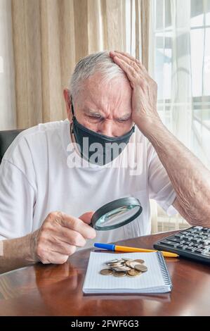 Alter Mann in einer medizinischen Maske mit einer Lupe untersucht und zählt Pennies aus der Pensionskasse. Besorgter älterer Mann steckt Geld in eine Sparschwein. ich Stockfoto