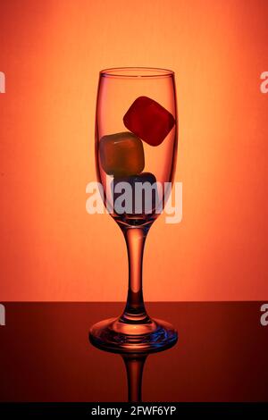 Sektglas (Flöte) mit farbigen Eiswürfeln darin auf einem reflektierenden Tisch mit orangefarbenem Hintergrund. Stockfoto