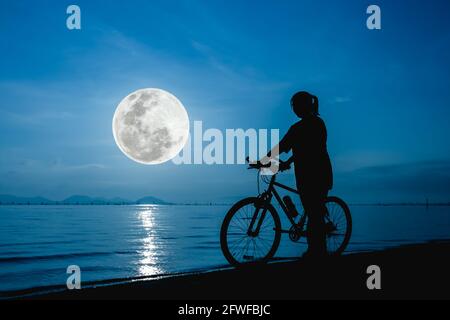 Silhouette eines gesunden Biker-Mädchens, das die Aussicht am Meer genießt, schöner Vollmond auf blauem Himmel Hintergrund. Spiegelung des Mondes im Wasser. Im Freien. Der Stockfoto