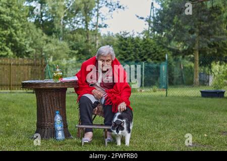 Ältere Frau, die draußen auf dem Pensionat sitzt, in rotem Poncho mit schwarz-weißem Katzenhüter Stockfoto