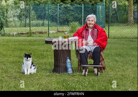 Ältere Frau, die draußen auf dem Pensionat sitzt, in rotem Poncho mit schwarz-weißem Katzenhüter Stockfoto