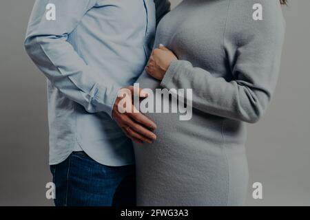 Die Schwangere trägt Kleiderposen in der Nähe des Mannes, der ihren großen Bauch berührt. Zukünftige Mutter und Vater warten auf ein neues Familienmitglied Stockfoto