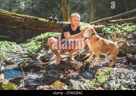 Ein lächelnder Mann mittleren Alters streichelt seinen Beagle-Hund in der Nähe des Bergwaldes, während er auf Wäschestauken und Trekkingstiefel wartet. Aktive Menschen trave Stockfoto