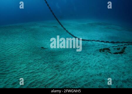 Unterwasserfoto einer schweren Stahlankerkette, die von der Wasseroberfläche herabhängt und auf weißem Sandboden liegt, dunkelblaues Wasser tiefer im tropischen Meer Stockfoto