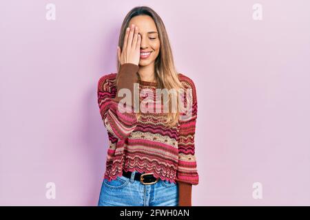 Schöne hispanische Frau mit Hippie-Pullover, der ein Auge mit der Hand bedeckt, selbstbewusstes Lächeln im Gesicht und überraschende Emotionen. Stockfoto