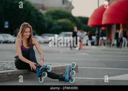 Zufrieden dunkelhaarige Europäerin setzt auf Inline-Skates gehen Rollerblading Posen vor verschwommenem Stadthintergrund hält fit verbringt frei Aktive Zeit Stockfoto