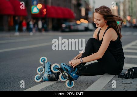 Die seitliche Aufnahme der aktiven Frau sitzt auf der Straße und verstellt Rollerblades Bereitet sich auf Skaten legt auf Inline-Skates hat Haare schweben Am Wind genießt das Wochenende Stockfoto