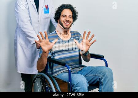 Gutaussehender hispanischer Mann, der auf dem Rollstuhl sitzt und einen Halsband trägt und mit den zehn Fingern nach oben zeigt, während er selbstbewusst und glücklich lächelt. Stockfoto
