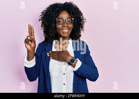 Die junge afroamerikanische Frau trägt Geschäftskleidung und eine Brille, die lächelnd mit der Hand auf der Brust und den Fingern nach oben flucht und einen Treueversprechen-Eid ablegt Stockfoto