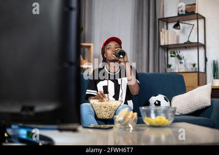 afroamerikanische Frau mit roten Mützchen trinkt Bier und isst Snacks, während sie sich ein Fußballspiel im Fernsehen anschaut. Konzept von Unterhaltung, Fan und Sport. Stockfoto