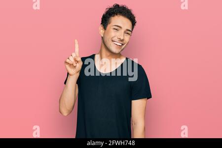 Ein junger, hübscher Mann, der ein legeres schwarzes T-Shirt trägt und mit der Fingernummer 1 nach oben zeigt und dabei selbstbewusst und glücklich lächelt. Stockfoto