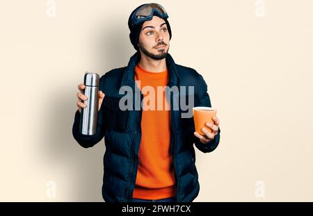 Junger hispanischer Mann mit Bart, der Schneekleidung trägt, und einer Himmelsbrille, die Kaffee hält und mit einem Lächeln auf die Seite blickt und dabei denkt. Stockfoto