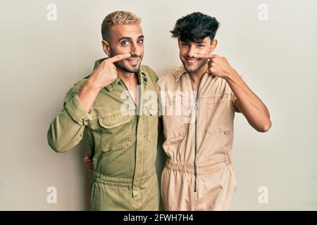 Homosexuelles, schwules Paar, das zusammen in einem lässigen Jumpsuit steht, der mit Finger auf Gesicht und Nase zeigt und fröhlich lächelt. Schönheitskonzept Stockfoto