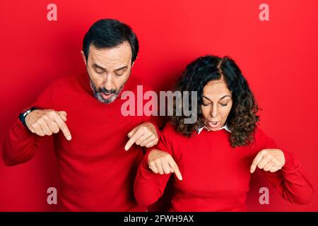 Ein Paar hispanischer Frauen und Männer mittleren Alters umarmte und stand zusammen und zeigte mit Fingern nach unten, die Werbung zeigten, überraschte das Gesicht und öffnete Mo Stockfoto