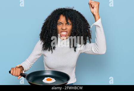 Junge afroamerikanische Mädchen hält Pfanne mit Spiegelei verärgert und frustriert schreiend vor Wut, schreiend verrückt vor Wut und die Hand angehoben Stockfoto