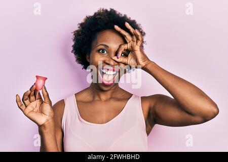 Junge afroamerikanische Frau hält Menstruationstasse lächelnd glücklich tun ok Schild mit Hand auf Auge durch die Finger schauen Stockfoto