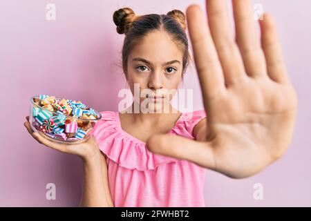 Schöne Brünette kleines Mädchen hält Glas voller Zucker Süßigkeiten mit offener Hand tun Stop-Zeichen mit ernsthaften und selbstbewussten Ausdruck, Verteidigung Geste Stockfoto