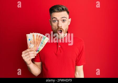 Junger Rotschopf mit philippinischen Peso-Banknoten erschrocken und erstaunt mit offenem Mund vor einem überraschenden, ungläubigen Gesicht Stockfoto