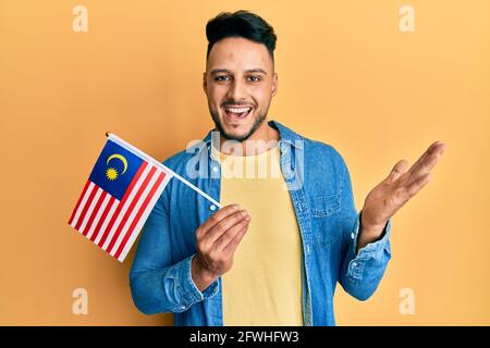 Junger araber, der die malaysische Flagge hält, feiert Erfolge mit Freude Lächeln und Ausdruck des Gewinners mit erhobener Hand Stockfoto