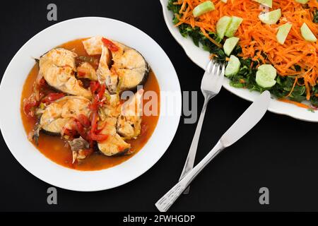 Gedämpfter Seebarsch in Sauce auf Teller mit grünem Salat auf schwarzem Hintergrund serviert. Stockfoto