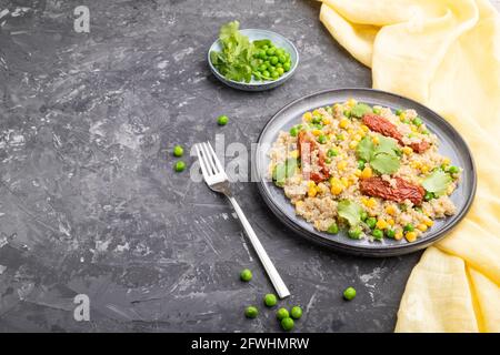 Quinoa-Haferbrei mit grüner Erbse, Mais und getrockneten Tomaten auf Keramikplatte auf grauem Betongrund und gelbem Textil. Seitenansicht, Kopierbereich. Stockfoto