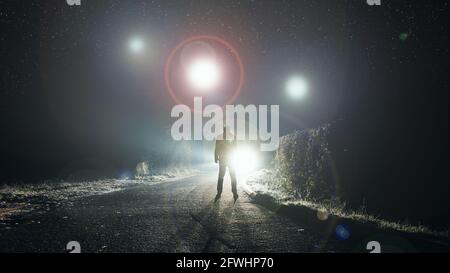 UFO-Konzept. Glühende Kugeln, die nachts über einer nebligen Straße schweben. Mit einer Silhouettenfigur, die auf die Lichter schaut. Stockfoto