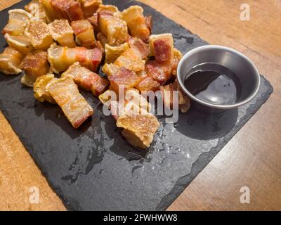 Nahaufnahme eines Tellers gebratener Torreznos oder gebratener Raschers auf einem schwarzen Schieferteller und eines Glases mit Grillsoße oder Sojasauce auf einem Holztisch, spanisch t Stockfoto