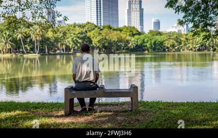 Ein nicht identifizierter alter Mann sitzt allein im Lumpini Park, Rückansicht eines mageren alten Mannes, der auf einem Steinsitz sitzt, einem großen Teich i Stockfoto
