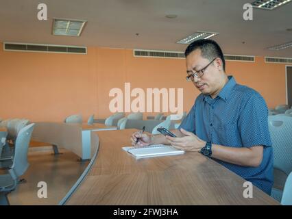 Ein Mann mittleren Alters aus Asien schaute auf ein Smartphone, hielt den Stift fest und öffnete das Notebook auf dem Schreibtisch und saß allein im Tagungsraum. Leerer Platz für Kopie an Stockfoto