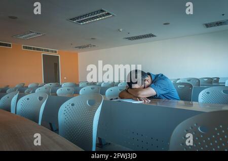Ein asiatischer Mann mittleren Alters schlaft allein auf dem langen Schreibtisch im Klassenzimmer, perspektivischer Blick, der Rest der Stühle ist leer. Stockfoto