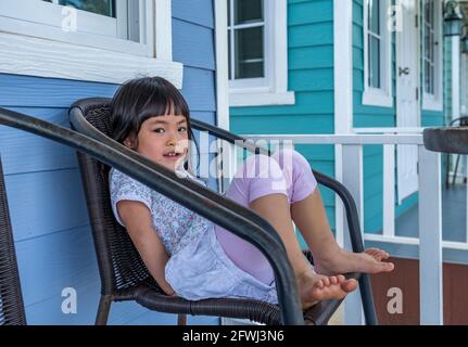 Portrait schönes asiatisches kleines Mädchen, das auf einem Stuhl vor der Holzwand des Resorthauses sitzt, Augen auf die Kamera schauen, lächelndes Gesicht, schwarzes l Stockfoto