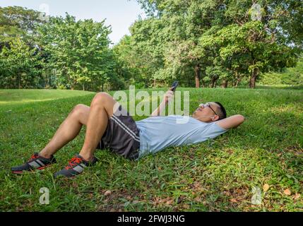 Der asiatische Jogger mittleren Alters legte sich in einem öffentlichen Park auf den Rasen und benutzte ein Mobiltelefon mit Kopfhörer. Stockfoto