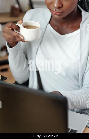 Zugeschnittenes Bild einer jungen Frau mit vollen Lippen Trinkbecher Von Kaffee und Arbeiten auf Laptop Stockfoto