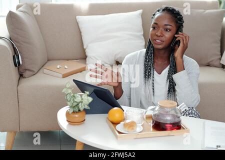 Seriöse Frau, die vor dem Tisch auf dem Boden sitzt Mit Teekannen und Tablet-Computer und Telefonieren mit Kollege Stockfoto