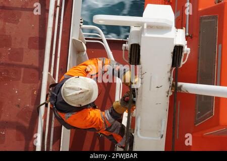 Nahaufnahme eines Crew-Mitglieds mit orangefarbener Jacke und weißem Helm, der mit einem Gurt am Metallgeländer befestigt ist. Stockfoto