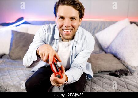 Nahaufnahme attraktiver junger Mann, hält einen Joy Stick und spielt Videospiele im Fernsehen, sitzt zu Hause auf einem gemütlichen Sofa Neon, erfolgreich, gewinnt er ein verrücktes Auto Stockfoto