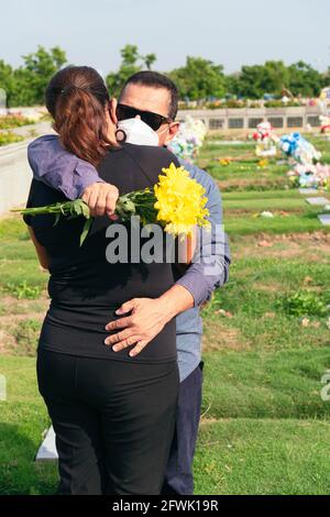 Reifes Paar mit Schutzmaske auf dem Friedhof umarmt während der Beerdigung. Tod, Friedhof, Verlust von Angehörigen aufgrund einer Pandemie. Stockfoto