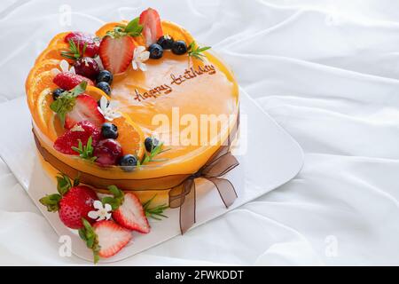 Orangenkuchen mit glücklichem Geburtstag und gekrönt mit Orange, Erdbeere, Heidelbeere auf weißem Stoffhintergrund, Kopierraum und Dessertkonzept Stockfoto