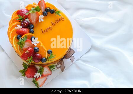 Orangenkuchen mit glücklichem Geburtstag und gekrönt mit Orange, Erdbeere, Heidelbeere auf weißem Stoffhintergrund, Kopierraum und Dessertkonzept Stockfoto