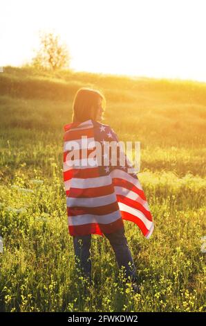Rückansicht einer jungen Frau, die bei Sonnenuntergang die US-Nationalflagge auf einem Feld hält. Unabhängigkeitstag Amerikas. Konzept der amerikanischen Freiheit