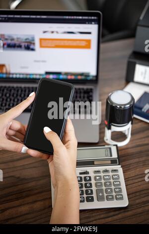 Smartphone in Frauenhand am Schreibtisch vor dem Hintergrund eines Rechners und eines Laptops. Stockfoto