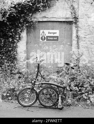 Mai 2021 - Brompton Faltrad vor einer bewachsenen Tür zu einem leeren Cottage-Gebäude im ländlichen Somerset, in der Nähe des Dorfes Wedmore. Stockfoto