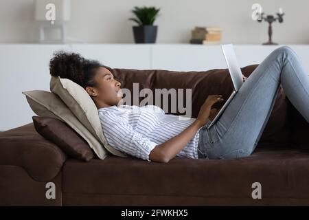 Entspannte, sorglose junge afrikanische Frau, die am Computer auf dem Sofa lag. Stockfoto