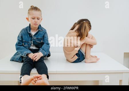 Traurige kleine Geschwister sitzen auf einem Tisch. Junge hat Mädchen beleidigt. Er ist traurig und schaut nach unten, unzufrieden mit sich selbst und dem Ausgang. Sie hat sich den Rücken gekehrt, Stockfoto