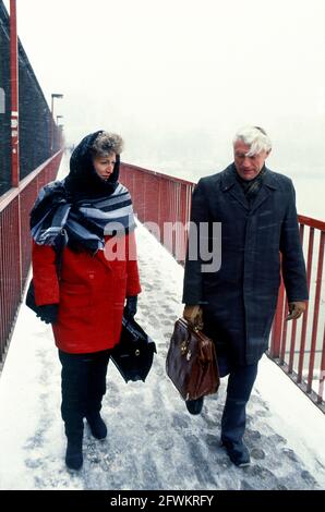 Zwei Personen, die die Themse, London, auf der Fußgängerbrücke entlang der Hungerford Eisenbahnbrücke (seitdem ersetzt) überqueren, bei fallendem Schnee Stockfoto