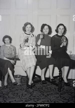 1957, historisch, auf einer Firmenfeier oder -Veranstaltung, in einem Veranstaltungsraum im Victoria Hotel, Leeds, drei junge Damen, die auf einem Tisch sitzen, zwei mit einem Bier für die Kamera, England, Großbritannien. Eine Frau, die neben ihnen auf einem Stuhl sitzt und eine Zigarette raucht, hat ebenfalls ein Pint Bier in der Hand. Stockfoto