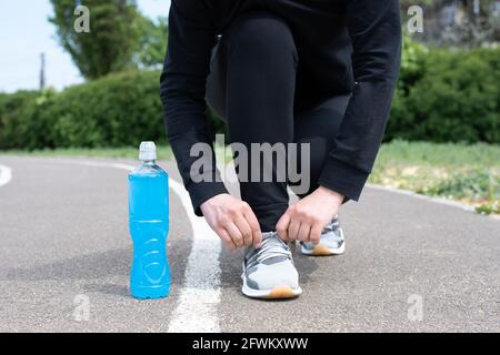 Eine blaue Plastikflasche mit isotonischem Getränk und eine Sportlerin, die Schuhe auf Turnschuhen auf der Laufstrecke aus nächster Nähe bindet. Stockfoto