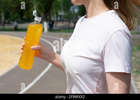 Sportlerin in weißem T-Shirt mit Flasche und orangefarbenem isotonischem Getränk auf Sportplatz, Sommer, Nahaufnahme. Stockfoto