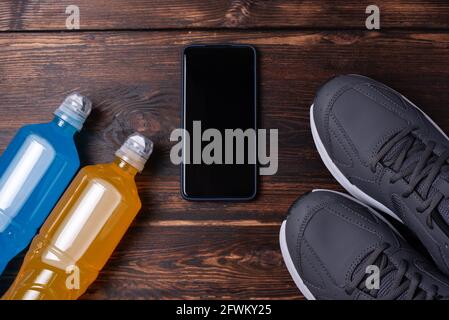 Graue Sneaker, Smartphone und zwei Flaschen isotonisches Getränk auf dunklem Holzhintergrund, Mockup, Sporttrainingskonzept, Nahaufnahme. Stockfoto